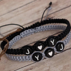 May include: Two friendship bracelets with black and gray cords. The bracelets have black beads with white letters spelling "JOY" and white hearts.