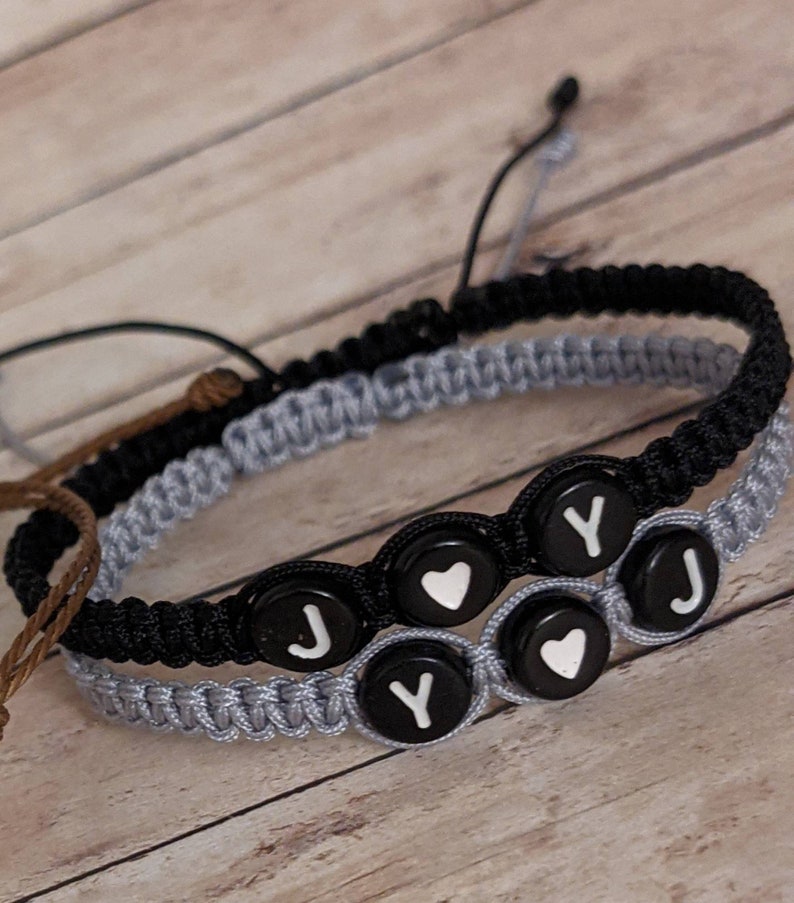 May include: Two friendship bracelets with black and gray cords. The bracelets have black beads with white letters spelling "JOY" and white hearts.