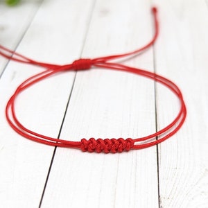 May include: A red string bracelet with a knotted design. The bracelet is made of two strands of string that are tied together in a series of knots.