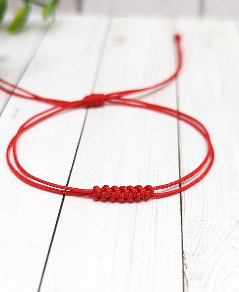 May include: A red string bracelet with a knotted design. The bracelet is made of two strands of string that are tied together in a series of knots.