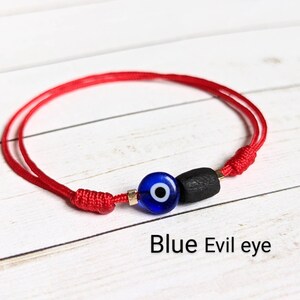 May include: Red string bracelet with a blue evil eye charm and a black bead. The bracelet is tied with a knot and the charm is in the center.