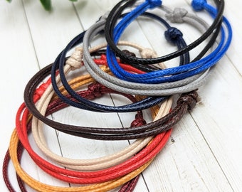 May include: A set of eight colorful braided bracelets in red, orange, yellow, beige, brown, navy blue, gray, and blue. The bracelets are made of thin cord and are tied with a knot at the end.