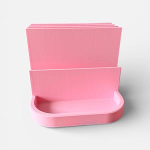May include: A pink desk organizer with a curved bottom and multiple vertical dividers. The organizer is made of a solid material and has a smooth, matte finish. The design is simple and modern, suitable for office or home use.
