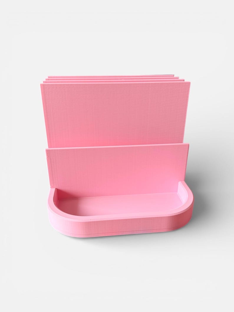 May include: A pink desk organizer with a curved bottom and multiple vertical dividers. The organizer is made of a solid material and has a smooth, matte finish. The design is simple and modern, suitable for office or home use.