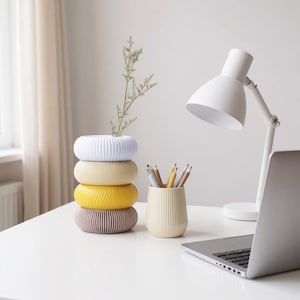 May include: A stack of four colorful, ribbed ceramic vases in white, beige, yellow, and mauve. A small beige vase holds pencils, and a white desk lamp and laptop are also on the white desk. A sprig of greenery is in the top vase.