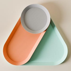 May include: A modern decorative tray set in a geometric design. The set includes a round gray tray resting on a larger tray with a peach-colored section and a mint-green section. The trays are made of a smooth material.