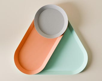 May include: A modern decorative tray set in a geometric design. The set includes a round gray tray resting on a larger tray with a peach-colored section and a mint-green section. The trays are made of a smooth material.