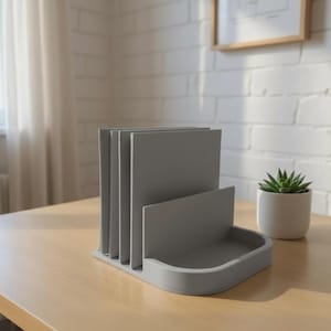 May include: A gray desk organizer with multiple vertical slots for papers and a small tray. The organizer is made of a solid material and sits on a light-colored wooden desk. A small potted succulent is to the right.