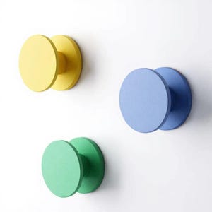 May include: Three circular wall hooks in solid matte colors. One hook is bright yellow, one is light blue, and one is a vibrant green. Each hook features a flat, round front face attached to a cylindrical base that extends from the wall surface.