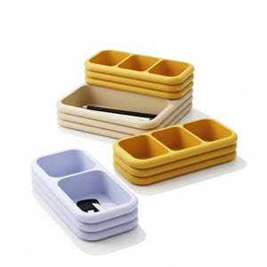 May include: A collection of colorful desk organizers in yellow, beige, and light blue. The organizers have multiple compartments and a stacked design. A pencil rests in one of the beige organizers. The organizers are made of a smooth material.