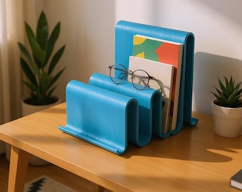 May include: A turquoise blue, wavy book organizer holds several books and a pair of round eyeglasses. The organizer is made of a ridged material and sits on a wooden table. A small potted plant is visible in the background.