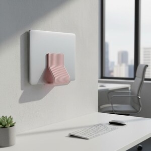 May include: A light pink, ribbed wall-mounted laptop holder attached to a white wall. The holder features a vertical, ridged design that securely supports a silver laptop in a closed, upright position, creating a clean and organized workspace.
