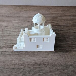 May include: A white 3D-printed model of a building with a domed tower, three windows, and a staircase. The miniature structure is set against a wooden surface.