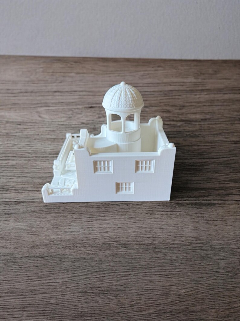 May include: A white 3D-printed model of a building with a domed tower, three windows, and a staircase. The miniature structure is set against a wooden surface.