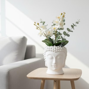 May include: A white ceramic Buddha head planter holding white orchid flowers and green eucalyptus leaves. The planter sits on a light wood side table. A gray sofa is in the background.