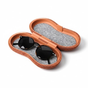 May include: An open, oval-shaped, terracotta-colored storage case with a ribbed exterior and a grey felt interior lining. Inside the case rests a pair of round-framed sunglasses with dark lenses and a dark tortoiseshell-patterned frame.