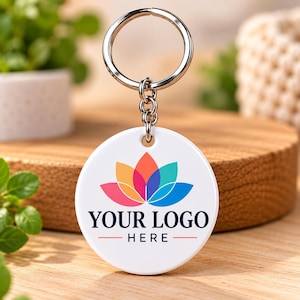 May include: A white circular keychain featuring a colorful lotus flower graphic in shades of pink, orange, and blue, with the black text "YOUR LOGO HERE" printed below the design, attached to a silver metal split ring and chain.
