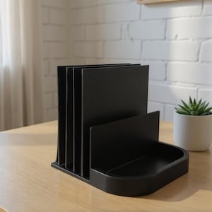 May include: A black desk organizer with multiple vertical slots for papers and a small tray. The organizer is made of a solid material and sits on a light-colored wooden desk. A small potted succulent is visible in the background.