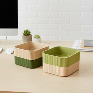 May include: Two square storage containers with ribbed sides. One is tan over green, the other is green over tan. The containers sit on a light wood surface with a white brick wall in the background. Small potted plants are also visible.