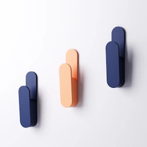May include: Three modern wall hooks in a row, each with a rounded rectangular shape. One is peach, and the other two are navy blue. The hooks are mounted on a white wall, providing a minimalist design.