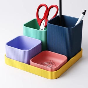 May include: A multi-coloured desk tidy set with a yellow base. It features teal, blue, and purple containers, a small pink tray with paper clips, red scissors, a black pen, and a white pen.