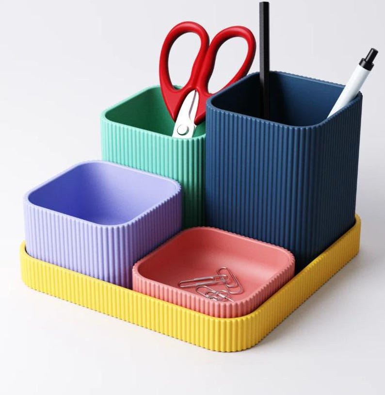 May include: A multi-coloured desk tidy set with a yellow base. It features teal, blue, and purple containers, a small pink tray with paper clips, red scissors, a black pen, and a white pen.