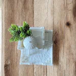 May include: A white, wall-mounted planter with a detailed architectural design, holding a small cluster of green artificial plants. The planter has a textured surface and is mounted on a wooden background.