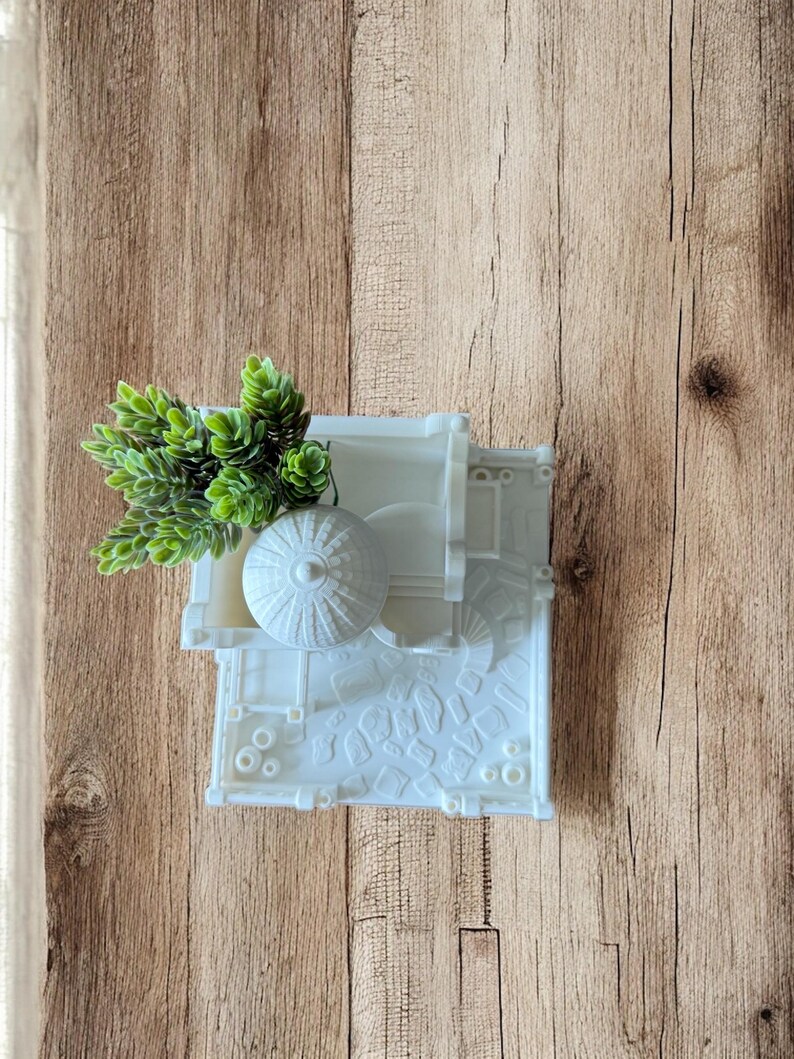 May include: A white, wall-mounted planter with a detailed architectural design, holding a small cluster of green artificial plants. The planter has a textured surface and is mounted on a wooden background.