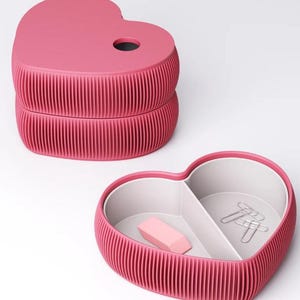 May include: A pink heart-shaped desk organizer with a ribbed exterior. The top section has a hole, and the bottom section is open, revealing a divided interior with a pink eraser and paper clips. The organizer is designed for office or desk use.