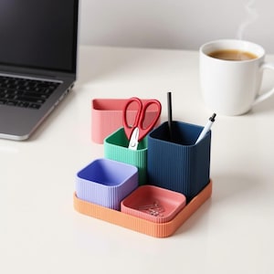 May include: A multi-compartment desk tidy in various colours. The set includes a coral tray, a small red dish, a purple square container, a green square container, a red rectangular container, and a navy blue square container. Scissors, pens, and paper clips are inside.