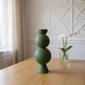 May include: A green ceramic vase with a stacked spherical design. The vase has a textured, ribbed surface and a small, rounded top. It stands on a wooden table, with a vase of white flowers in the background.