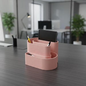 May include: A light pink desk organizer with a ribbed texture and three tiered compartments. The organizer holds a black spiral-bound notebook, a yellow pencil, and two pens, providing a structured space for office supplies on a dark surface.