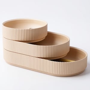 May include: A three-tiered, beige-colored serving tray. Each oval-shaped tier has a ribbed exterior and a smooth interior. The trays are stacked in a cascading design, offering a stylish way to display items. The overall design is minimalist and modern.