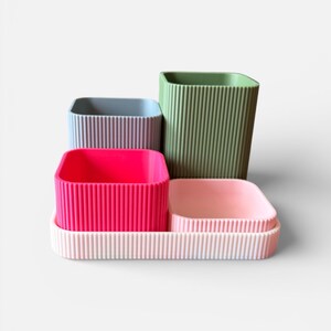 May include: A collection of four colourful, ribbed containers in shades of pink, green, blue, and a light pink tray. The containers are square or rectangular and vary in size, with vertical ribbing on the sides.