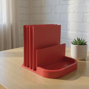 May include: A red desk organizer with multiple vertical slots for papers and a small tray. The organizer is made of a solid material and sits on a light-colored wooden surface. A small potted succulent is visible in the background.