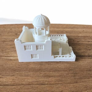 May include: A white miniature architectural model of a building with a domed gazebo, windows, and a balcony. The model is made of a solid white material and sits on a wooden surface.