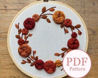 May include: Embroidered hoop art featuring a wreath of orange and red roses with green leaves. The hoop is made of wood and has a white fabric background. The text "PDF Pattern" is visible in the lower right corner of the image.