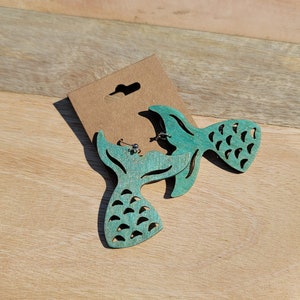 May include: A pair of teal blue mermaid tail earrings made of wood. The earrings are on a brown card.