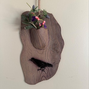 May include: A brown ceramic wall hanging with a carved wood grain design. It features a small planter with a sprig of flowers and a black silhouette of a crow.
