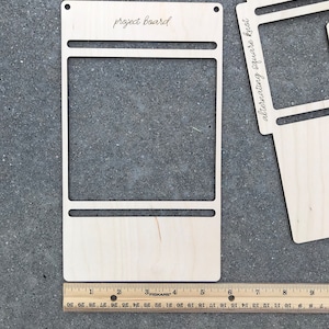 May include: A light-colored wooden project board with a rectangular cutout and horizontal slots, featuring the text 'project board' at the top. A wooden Fiskars ruler is positioned at the base, showing the board is approximately 9 inches wide.