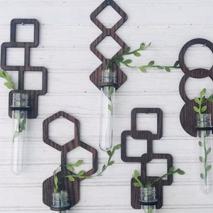 May include: Wooden wall-mounted vases in various geometric shapes, including squares, diamonds, and circles. Each vase holds a clear glass tube with greenery. The vases are stained a dark brown and mounted on a white, wood-paneled wall.