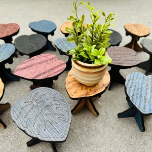 May include: A collection of small wooden plant stands in various colors and designs, including a heart shape, a leaf shape, and a simple round shape. A small potted plant sits on one of the stands.
