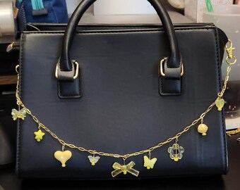 Puede incluir: Bolso de mano negro con asa superior y herrajes dorados. Una cadena dorada con abalorios amarillos, incluyendo corazones, estrellas y mariposas, cuelga en la parte delantera. El bolso tiene forma rectangular.