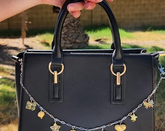May include: Black handbag with a silver chain and decorative charms. The charms include butterflies, stars, hearts, and bows in yellow and clear colors. The bag has a top handle and gold-colored hardware.