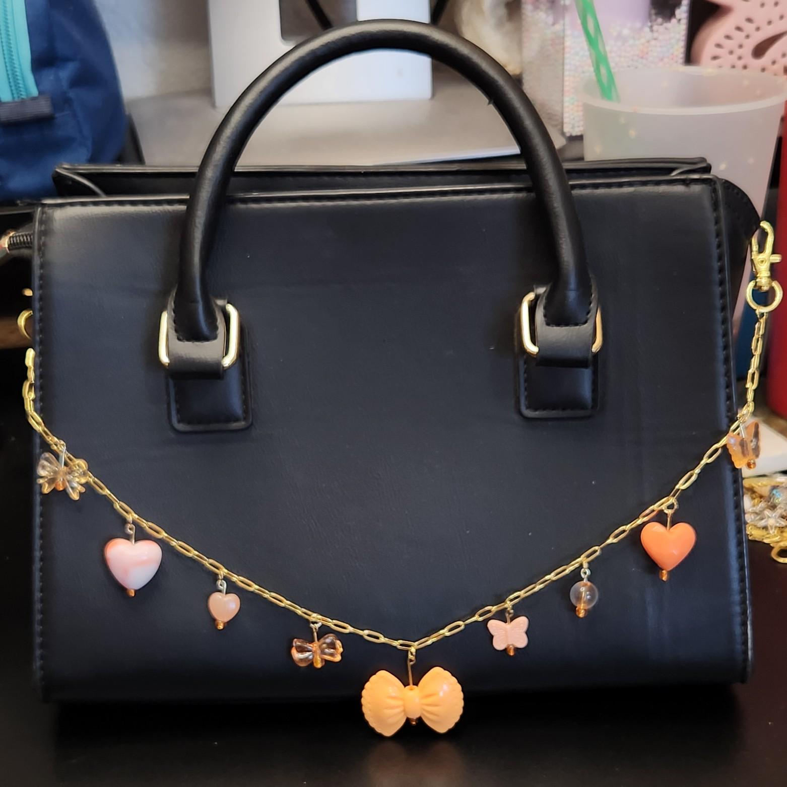 Puede incluir: Un bolso negro con asa superior y herrajes dorados. Una cadena dorada con abalorios naranjas y rosas, incluyendo corazones, mariposas y un lazo, cuelga en la parte delantera. El bolso tiene forma rectangular y un diseño estructurado.
