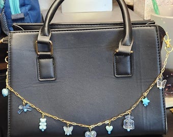 Puede incluir: Un bolso negro con una correa de cadena dorada adornada con abalorios azules. Los abalorios incluyen mariposas, corazones y un osito de peluche. El bolso tiene un asa superior y herrajes dorados.
