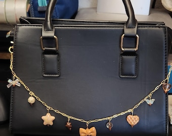 Puede incluir: Un bolso negro con una cadena dorada y abalorios decorativos. Los abalorios incluyen un lazo, estrellas, corazones y cuentas de varios colores. El bolso tiene forma rectangular con un asa superior.