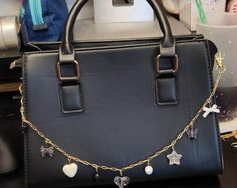 May include: Black handbag with a top handle and gold-colored hardware. A gold chain with charms, including a white heart, a clear heart, a star, and a bow, hangs from the bag.