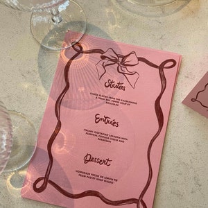Op de afbeelding: Een roze menu met een rood lintontwerp. Het menu bevat voorgerechten, hoofdgerechten en desserts. De tekst op het menu luidt: "Starters", "Kaasplateau van de Kaaskoning & Verse Paprika Soep van Boschaak", "Entrées", "Italiaanse vegetarische lasagne met pompoen, cottage cheese en Parmezaanse kaas", "Dessert", "Zelfgemaakte pecan- of citroenpastei van banketbakker Waldo".