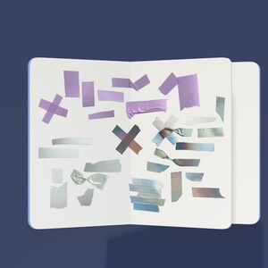 Op de afbeelding: Een open notitieboek met verschillende washi tapes in verschillende kleuren en patronen, waaronder paars, zilver en iriserend. De tapes zijn willekeurig gerangschikt op de witte pagina's.